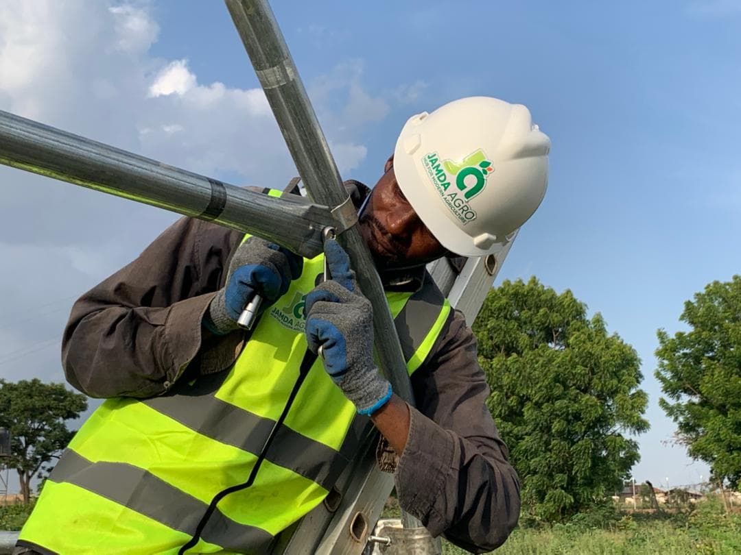 A Jamdaagro worker in a branded hard hat and high-visibility vest assembling the steel frame of a greenhouse
