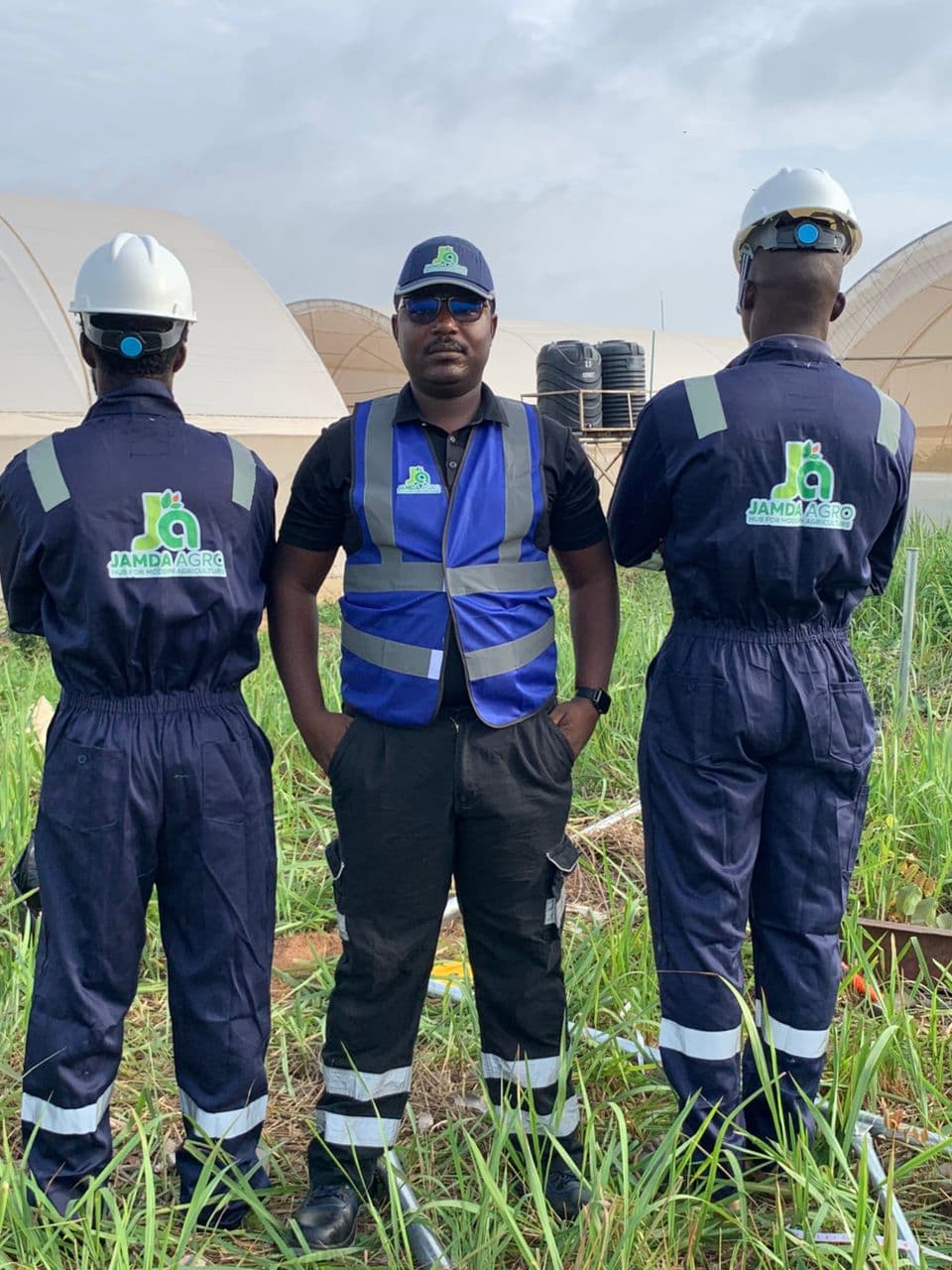 Three members of the Jamdaagro team in branded coveralls and a high-visibility vest standing in front of the greenhouse tunnels